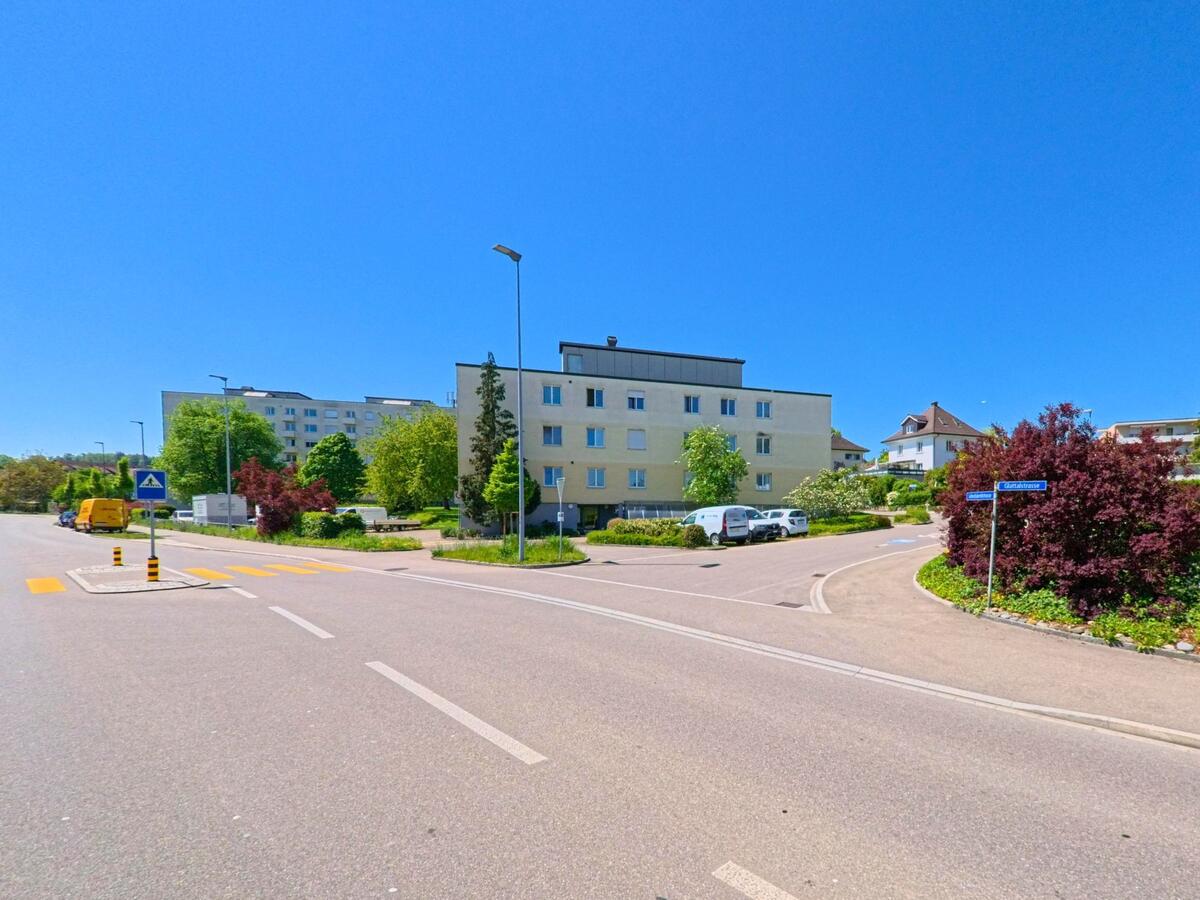 In Rümlang (8153, Kanton Zürich) verläuft eine breite Strasse mit Zebrastreifen durch ein ruhiges Wohngebiet mit gepflegten Mehrfamilienhäusern und farbenreicher Bepflanzung.