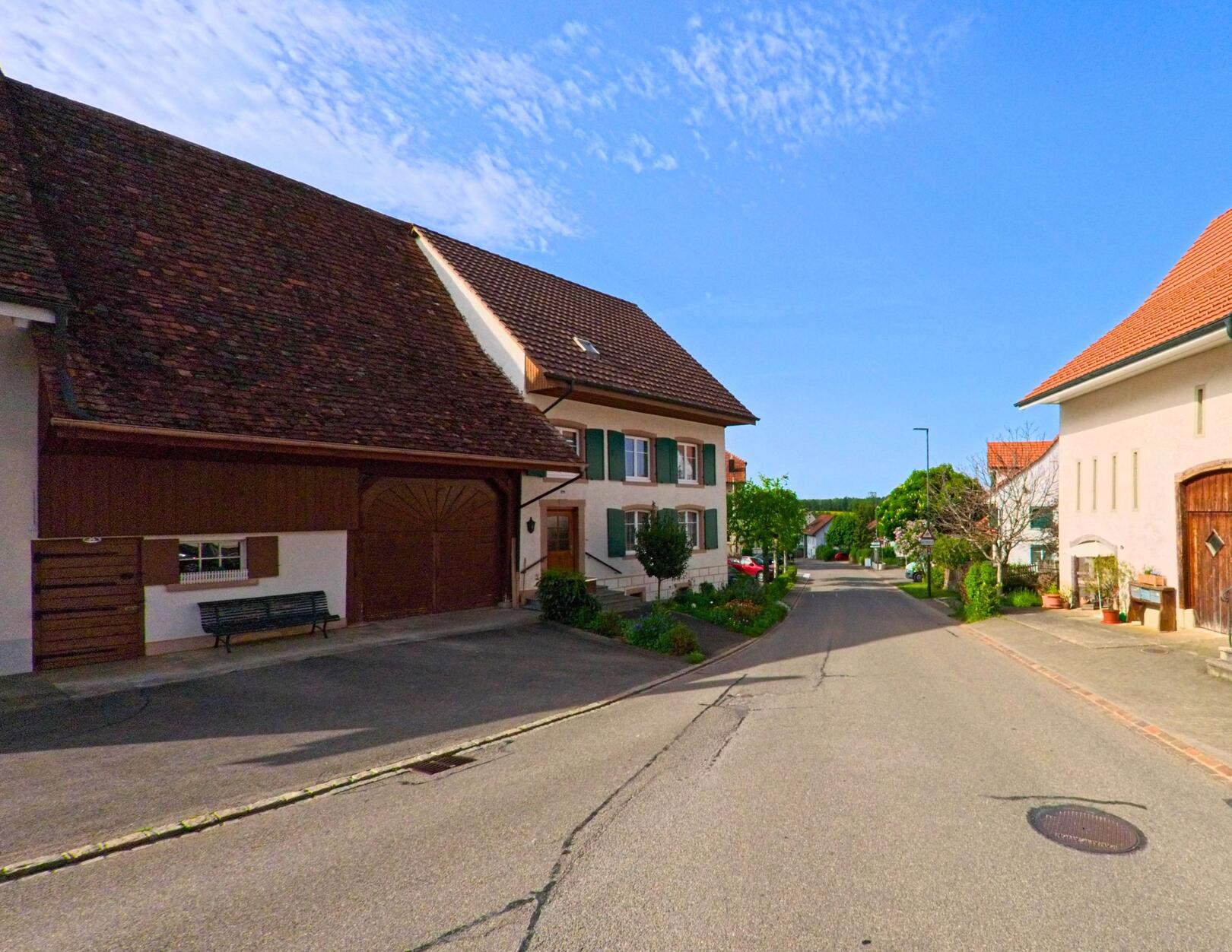 In Rünenberg (4497, Kanton Baselland) säumen gepflegte Bauernhäuser mit grünen Fensterläden und Scheunentoren eine ruhige Dorfstrasse.