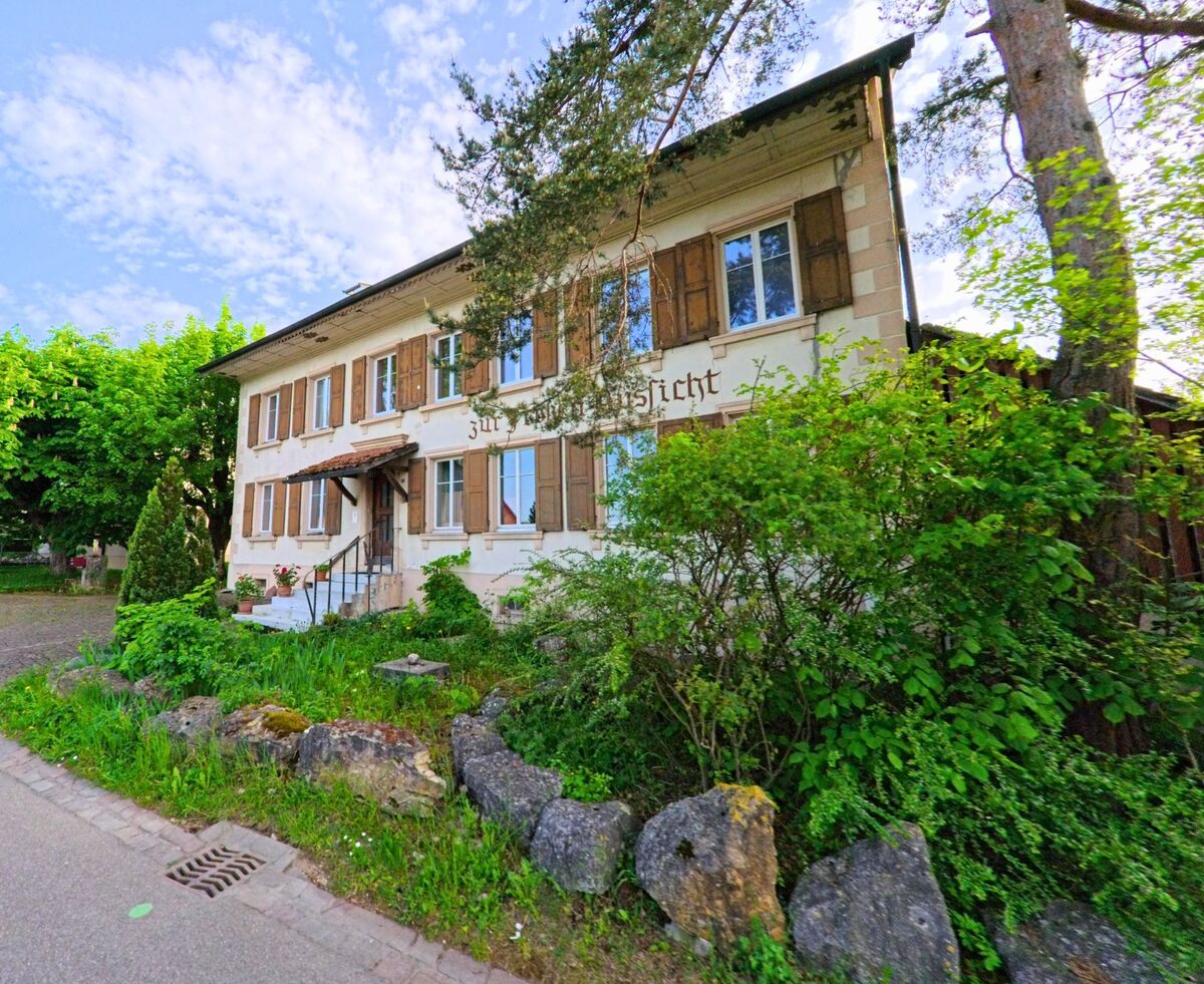 In Rünenberg (4497, Kanton Baselland) steht ein altes Gasthaus mit Holzläden und der Aufschrift „Zur Aussicht“ an einer leicht geschwungenen Strasse, umgeben von Bäumen und Sträuchern.