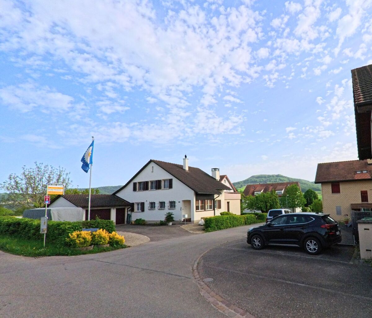 In Rünenberg (4497, Kanton Baselland) mündet eine Wohnstrasse in eine kleine Kreuzung, flankiert von Einfamilienhäusern, parkenden Autos und einer wehenden Gemeindeflagge.