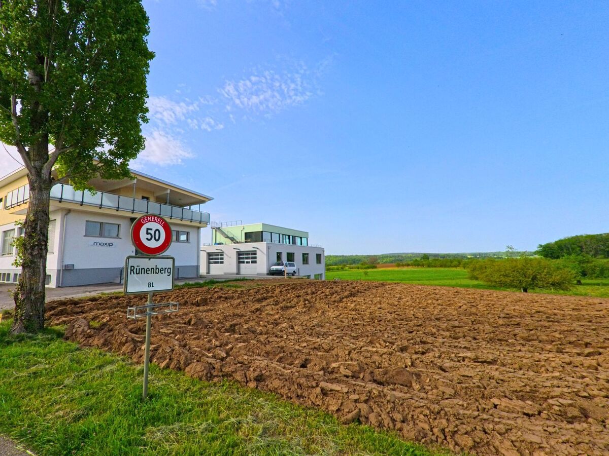 Am Ortseingang von Rünenberg (4497, Kanton Baselland) steht ein Geschwindigkeitsschild neben einem modernen Firmengebäude und frisch gepflügtem Ackerland.