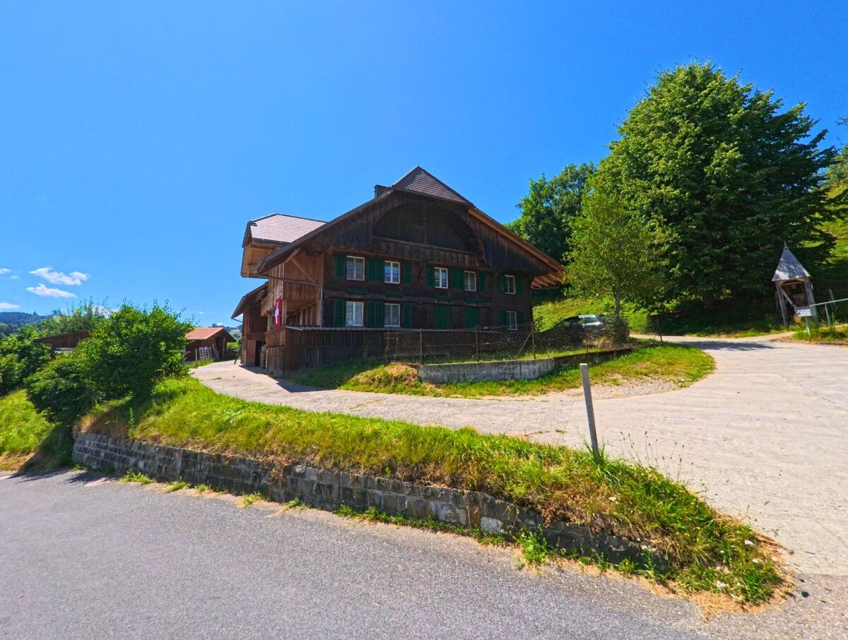 In Rüschegg (3153, Kanton Bern) steht ein traditionelles Bauernhaus mit Schweizer Flagge an einem Hang, daneben führt ein befestigter Weg zu einem Pavillon unter Bäumen.