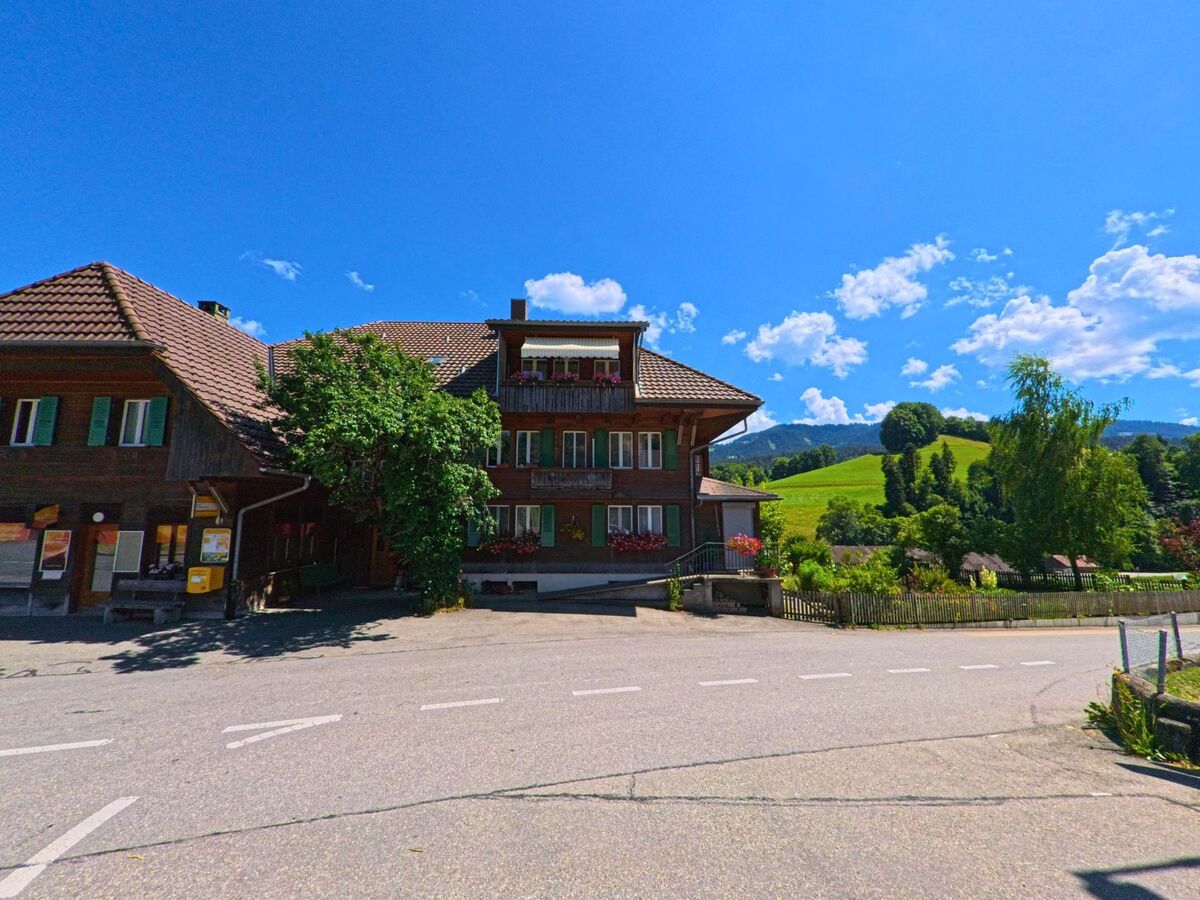 Vor der Kulisse grüner Hügel steht in Rüschegg (3153, Kanton Bern) ein traditionelles Wohnhaus mit roten Blumenbalkonen an einer leicht abschüssigen Strasse.