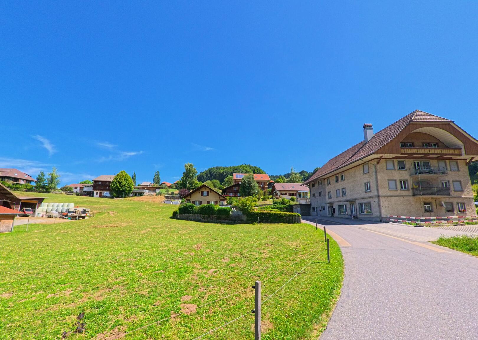 Am Rand einer grossen Wiese in Rüschegg (3153, Kanton Bern) stehen mehrere Wohnhäuser, darunter ein markantes Gebäude mit Walmdach und Balkon.