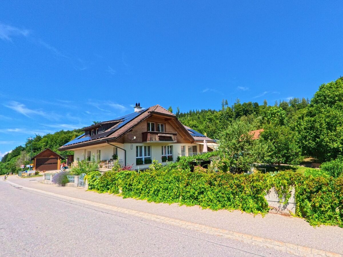 In Rüschegg (3154, Kanton Bern) steht ein Einfamilienhaus mit Solarpanels und gepflegtem Vorgarten an einer sonnigen Strasse, umgeben von dichter Vegetation.
