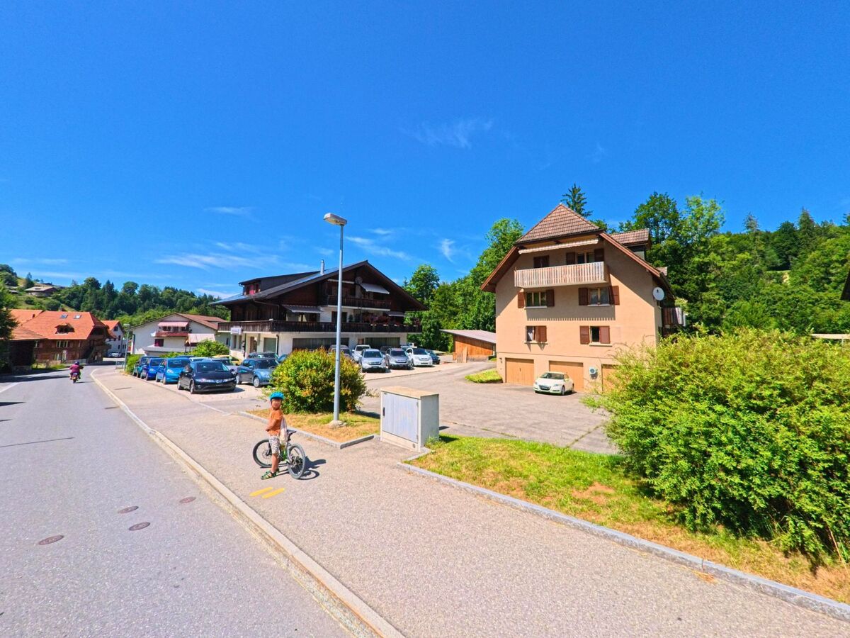 In einem Wohnquartier in Rüschegg (3154, Kanton Bern) steht ein Junge mit Fahrrad auf dem Trottoir, während sich im Hintergrund mehrere Einfamilienhäuser und geparkte Autos befinden.