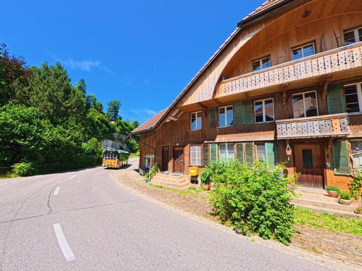 Ein gelber Postauto-Bus fährt eine kurvige Strasse entlang eines traditionellen Holzhauses mit grünen Fensterläden in Rüschegg (3154, Kanton Bern) hinauf.
