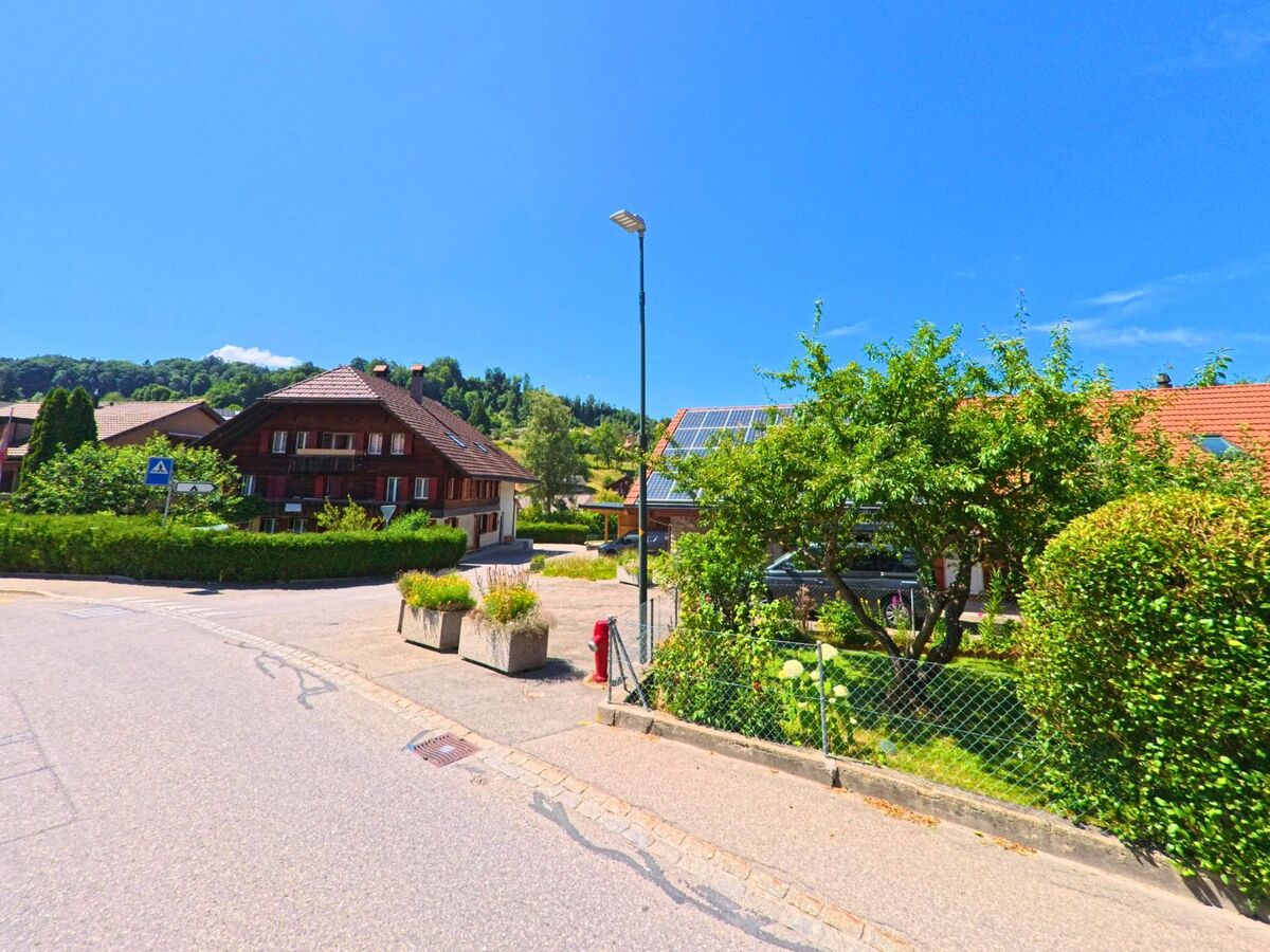 In einem sonnigen Wohngebiet von Rüschegg (3154, Kanton Bern) stehen ein traditionelles Berner Haus und ein Einfamilienhaus mit Solarpanels hinter gepflegten Gärten.