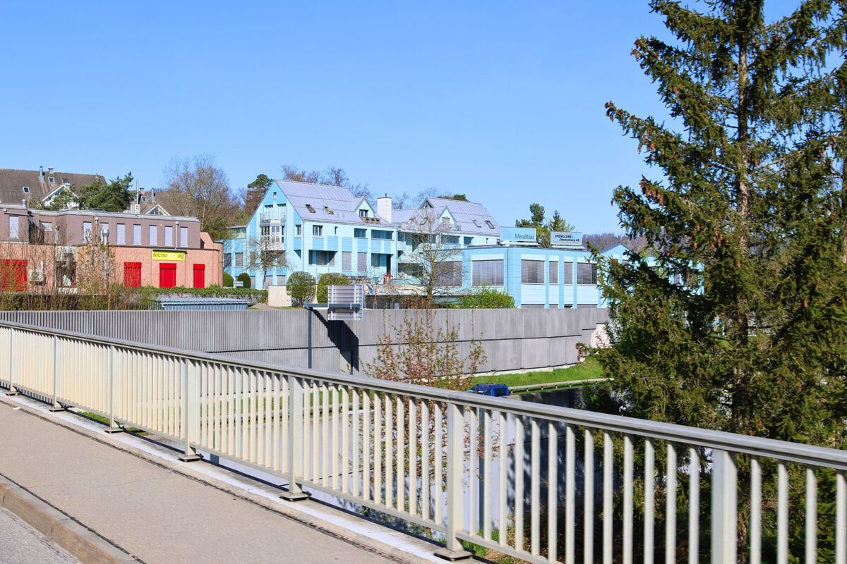 Mehrere farblich auffällige Gebäude mit Gewerbenutzung stehen in Rüschlikon im Kanton Zürich (PLZ 8803) oberhalb einer Lärmschutzwand, gesehen von einer Brücke mit Geländer.