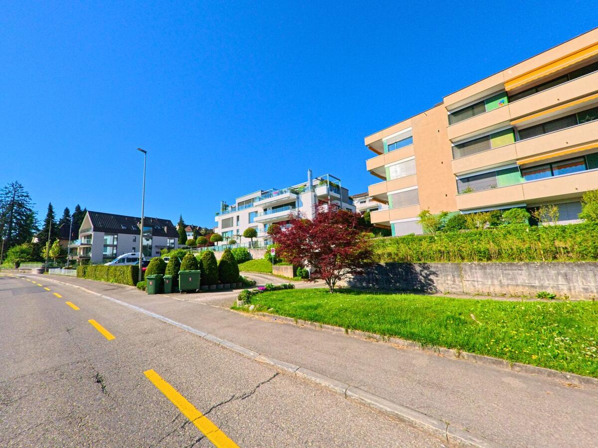 In Rüschlikon im Kanton Zürich (PLZ 8803) säumen moderne Mehrfamilienhäuser mit gepflegten Vorgärten eine sanft ansteigende Wohnstrasse unter klarem Himmel.