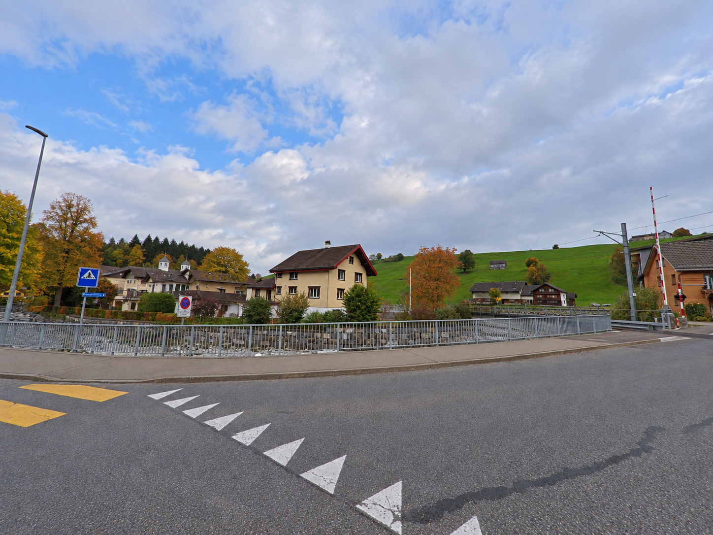An einer Strasse in Rüte(9050, Kanton Appenzell Innerrhoden) stehen Wohnhäuser entlang eines Bachs, während im Hintergrund grüne Hänge und ein Bahnübergang sichtbar sind.