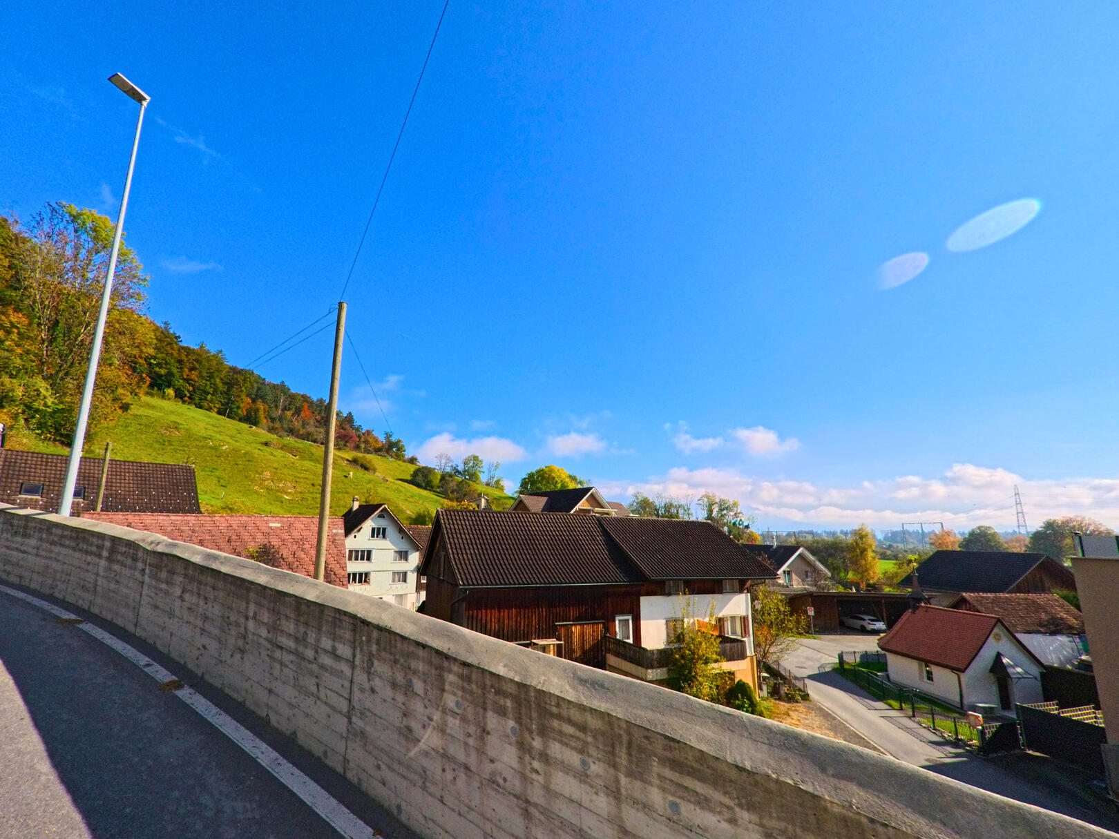 Entlang einer leicht erhöhten Strasse zeigt sich ein Wohnquartier mit Einfamilienhäusern in Ruethi (9464, Kanton St. Gallen) vor einem sonnigen Hang und blauem Himmel.