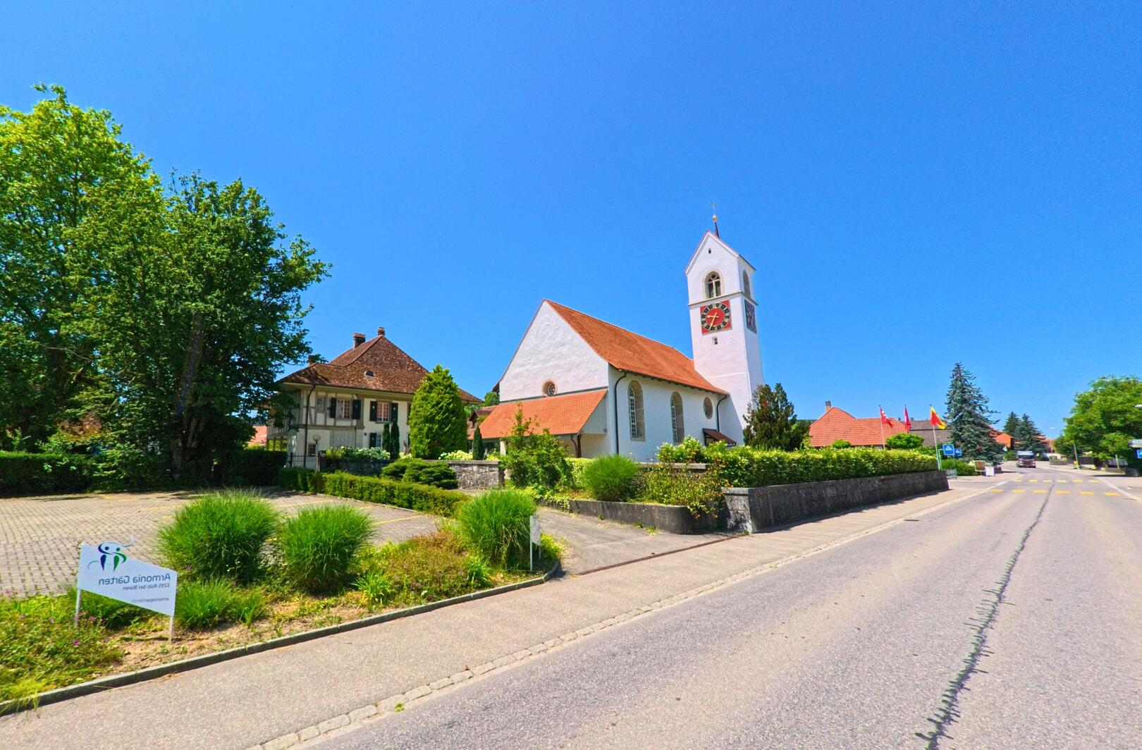 In Rüti bei Büren (3295, Kanton Bern) steht die weisse Kirche mit dem markanten roten Zifferblatt am ruhigen Strassenrand bei strahlend blauem Himmel.