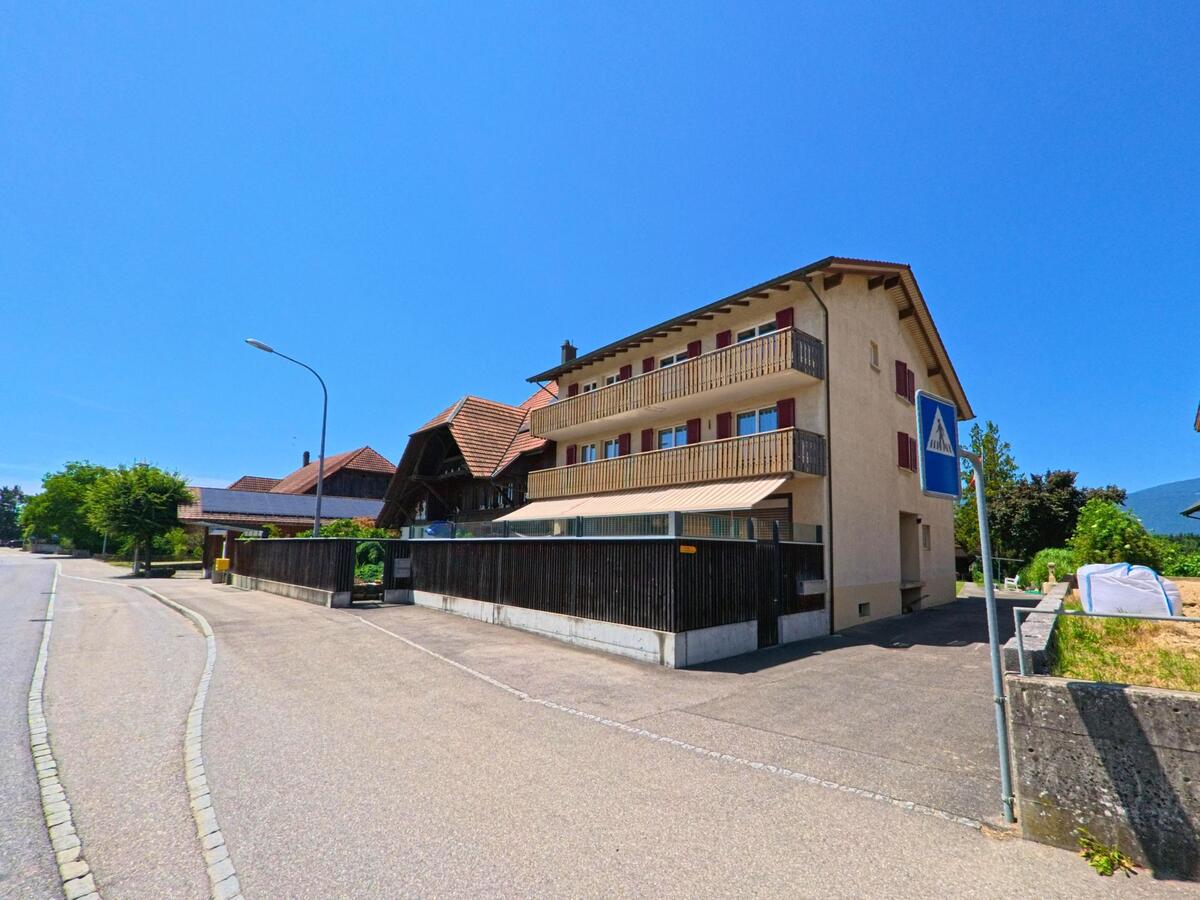 Ein Mehrfamilienhaus mit roten Fensterläden und Balkonen steht in Rüti bei Büren (3295, Kanton Bern) an einer ruhigen Strasse mit Zebrastreifen und Bushaltestelle.