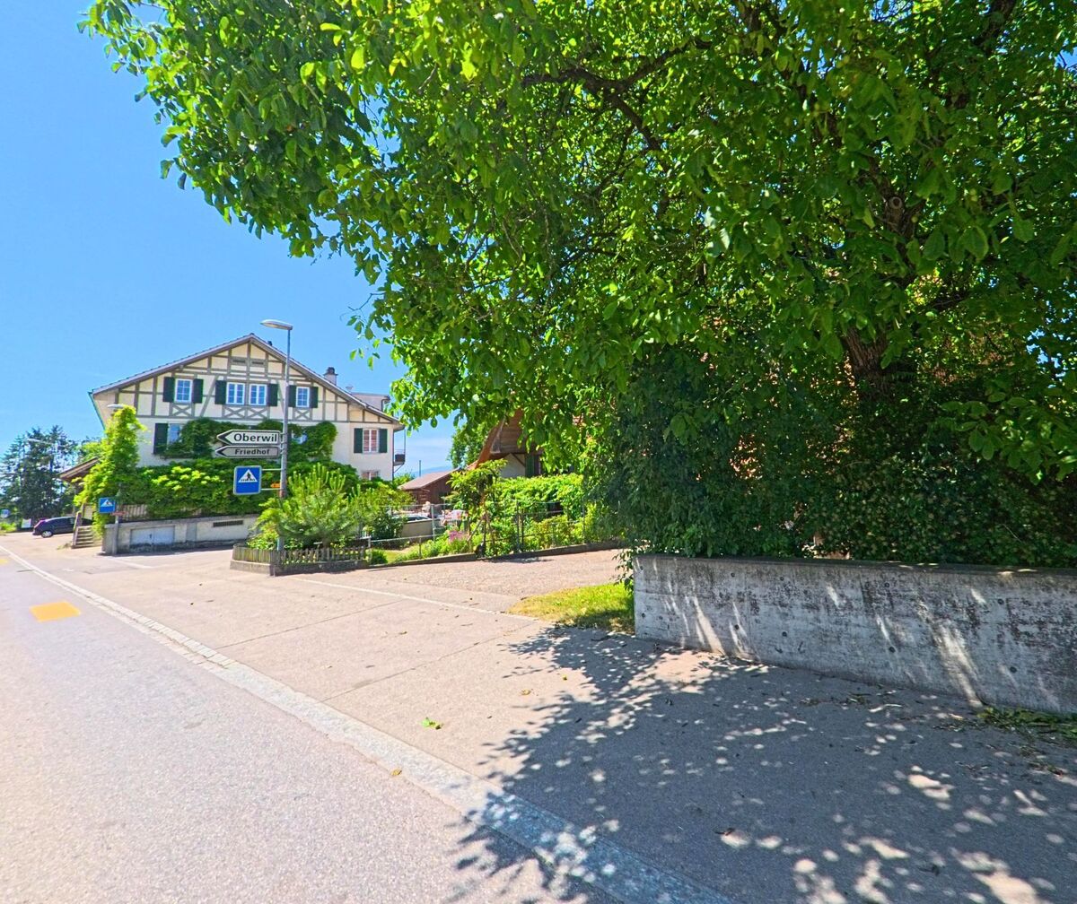 Ein Wegweiser in Rüti bei Büren (3295, Kanton Bern) zeigt in Richtung Oberwil und Friedhof, während im Hintergrund ein von Efeu bewachsenes Haus mit Fachwerkfassade zu sehen ist.