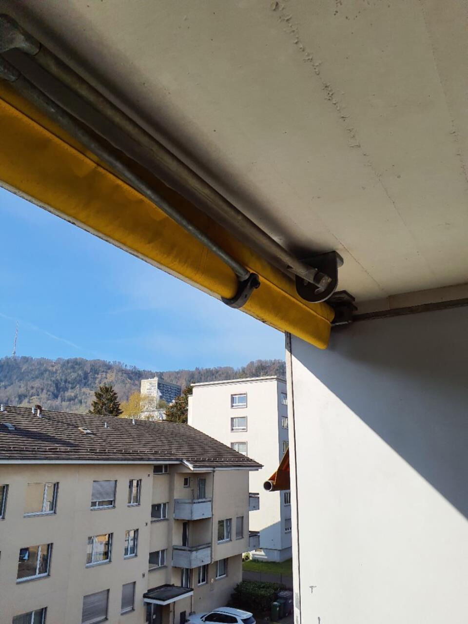 In Rüti (8632, Kanton Zürich) zeigt der Blick von einem Balkon mit gelber Markise auf mehrere helle Wohnhäuser vor einem bewaldeten Hügel im Hintergrund.