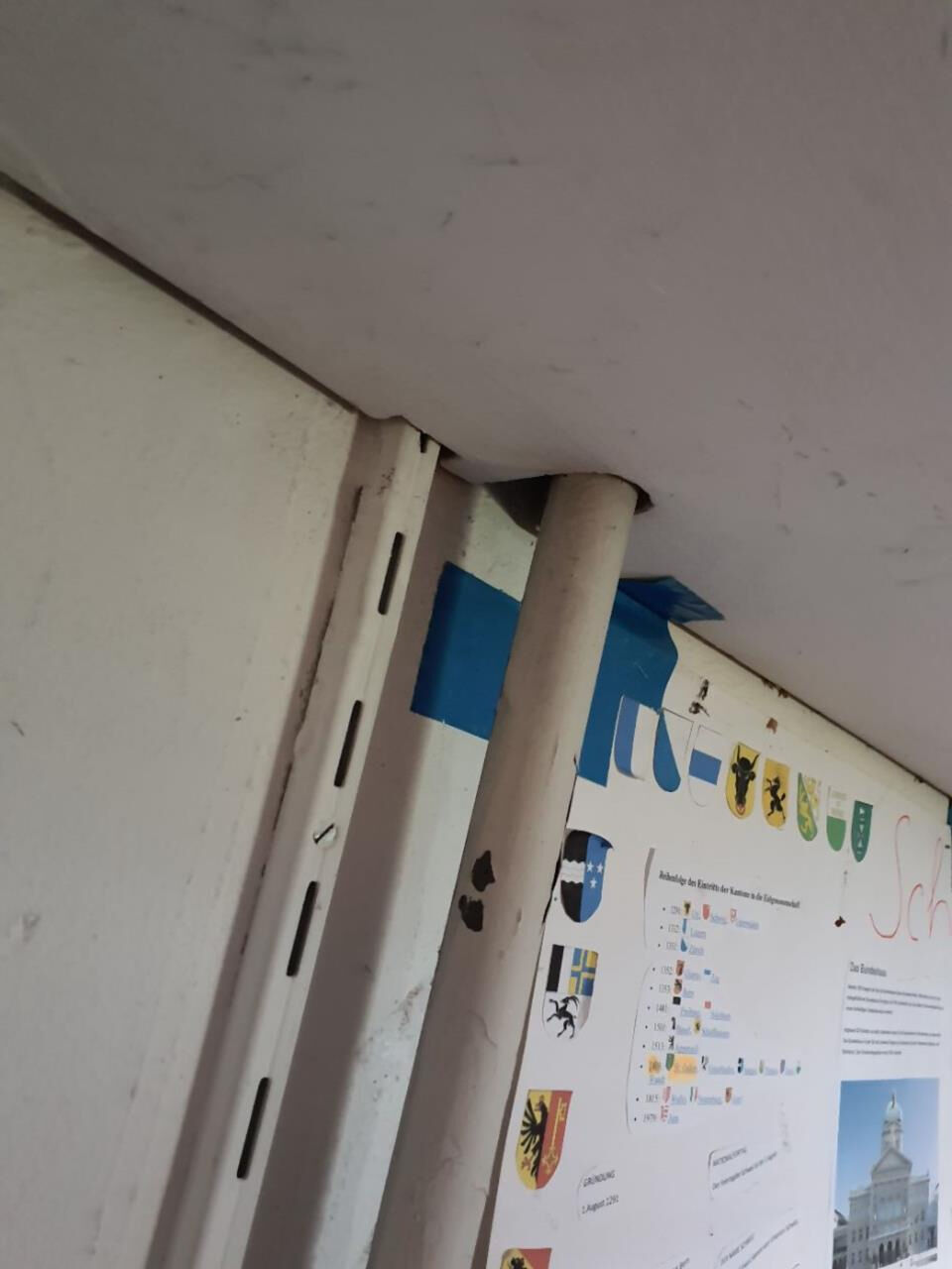  In Rüti (8632, Kanton Zürich) ist eine Infotafel mit Schweizer Kantonswappen an einer beschädigten Metallhalterung befestigt, wobei Klebeband und Roststellen sichtbar sind.