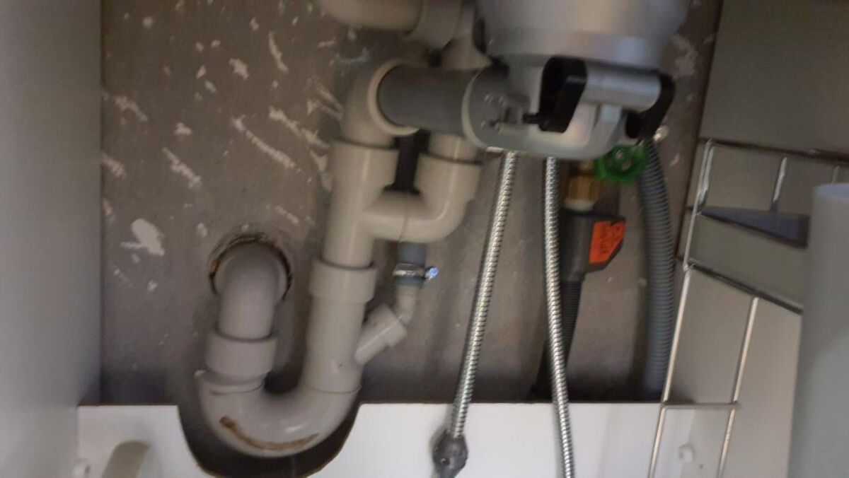 Unter einer Spüle in Rüti (8632, Kanton Zürich) verlaufen mehrere graue Abflussrohre vor einer unverputzten Wand, daneben sind Metallleitungen und ein Absperrventil sichtbar.