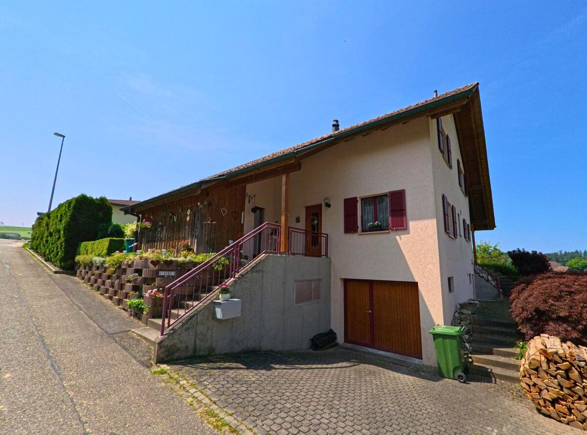 In Rütschelen (4933, Kanton Bern) steht ein Einfamilienhaus mit integriertem Garagentor und geschmücktem Eingangsbereich an einem leicht ansteigenden Strassenabschnitt.