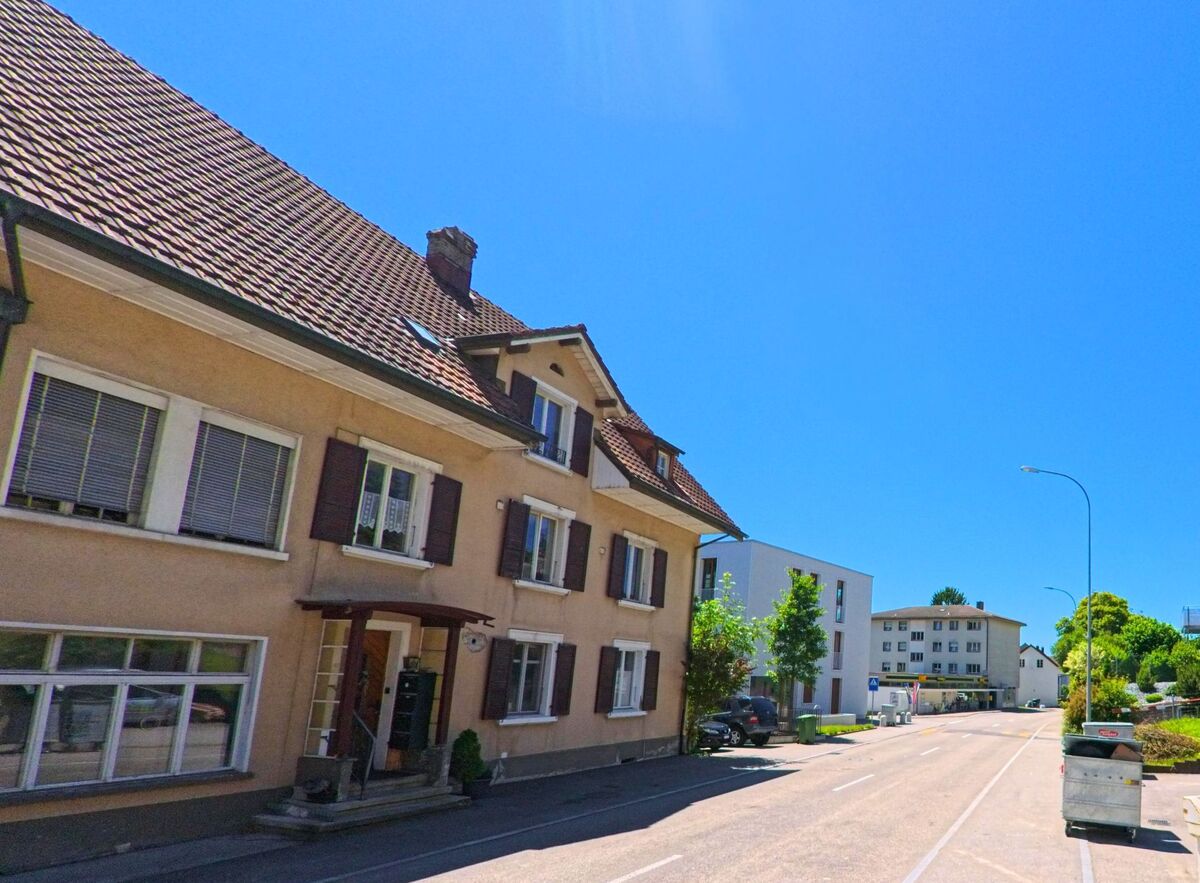 In Rüttenen (PLZ 4522, Kanton Solothurn) reiht sich ein älteres Wohnhaus mit Fensterläden an moderne Neubauten entlang einer sonnigen Strasse.