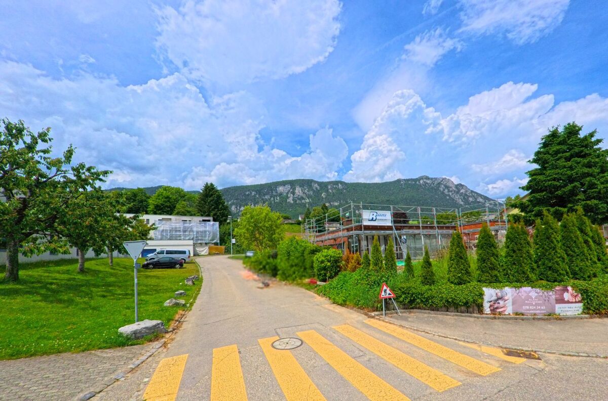In Rüttenen im Kanton Solothurn (PLZ 4522) führt ein Fussgängerstreifen zu einem kleinen Strässchen mit Baustelle, eingerahmt von Bäumen und dem Jura im Hintergrund.