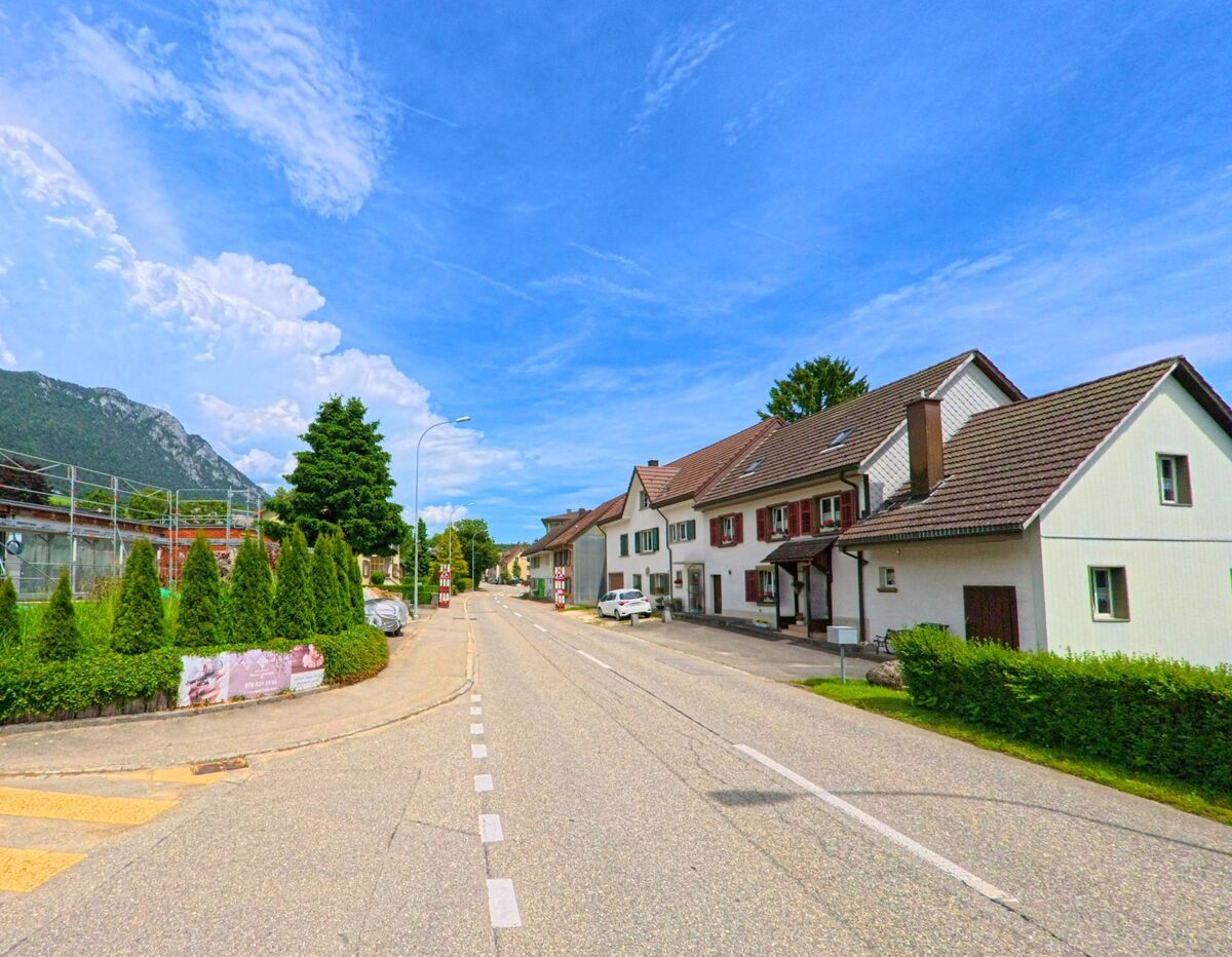  Entlang einer Hauptstrasse in Rüttenen (4522, Kanton Solothurn) reihen sich traditionelle Wohnhäuser mit Fensterläden, während im Hintergrund der bewaldete Jura sichtbar ist.