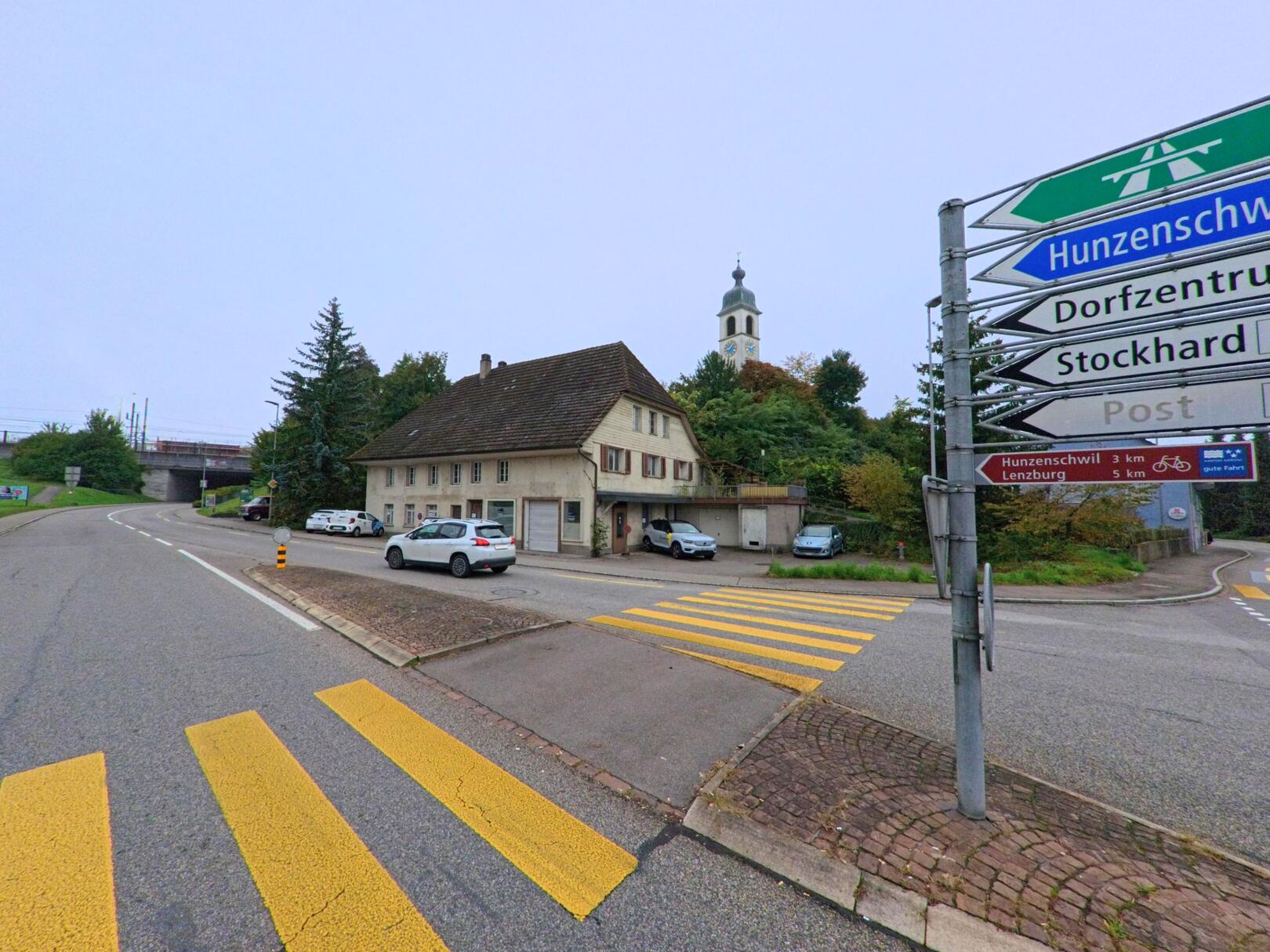 Ein Strassenzug in Rupperswil (5102, Kanton Aargau) mit Wegweisern nach Hunzenschwil und Lenzburg sowie Blick auf ein altes Wohnhaus und den Kirchturm im Hintergrund.