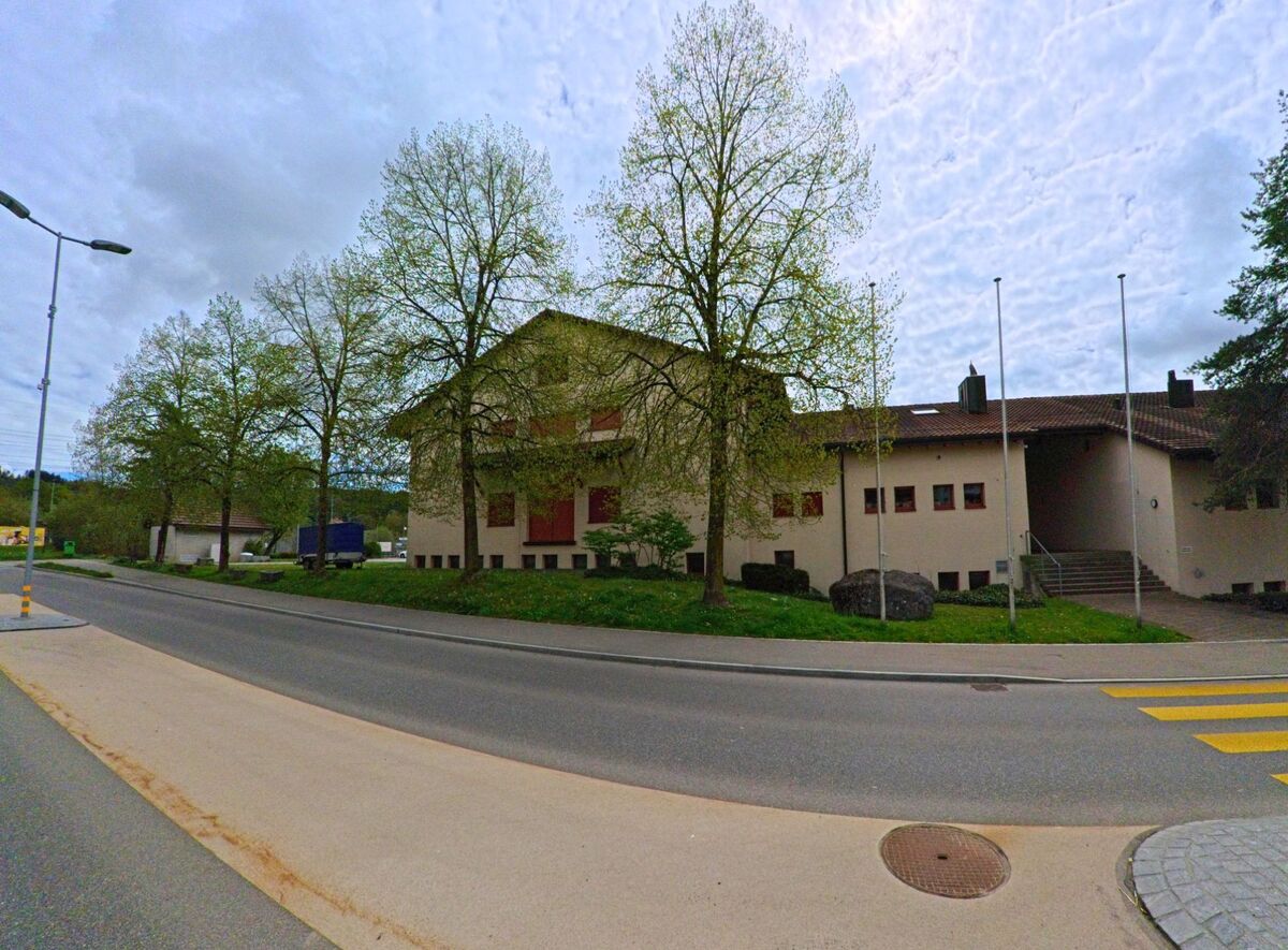 Gegenüber einer Strasse in Russikon (8332, Kanton Zürich) steht ein grösseres Gebäude mit roten Fensterläden hinter einer Reihe junger Laubbäume.