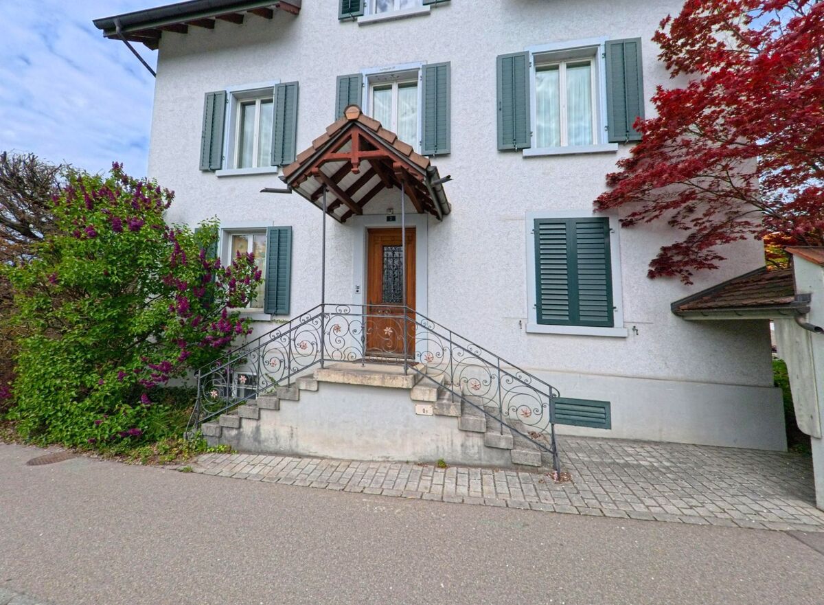 In Russikon (8332, Kanton Zürich) führt eine verzierte Steintreppe mit Metallgeländer zu einem Hauseingang mit Vordach, flankiert von blühenden Sträuchern.