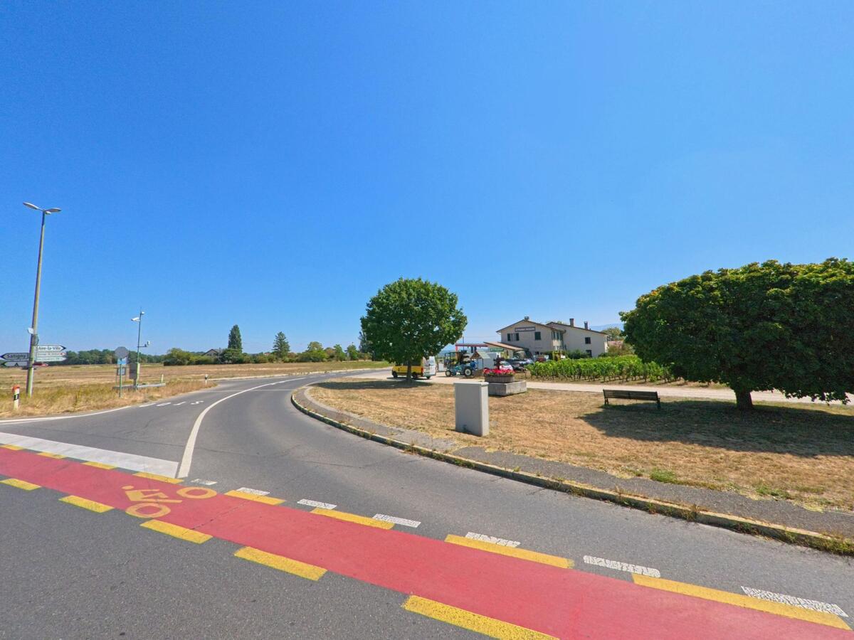 Am Ortseingang von Russin (1281, Kanton Genf) verläuft eine Landstrasse mit markiertem Radweg vorbei an Feldern und einem Wohnhaus.