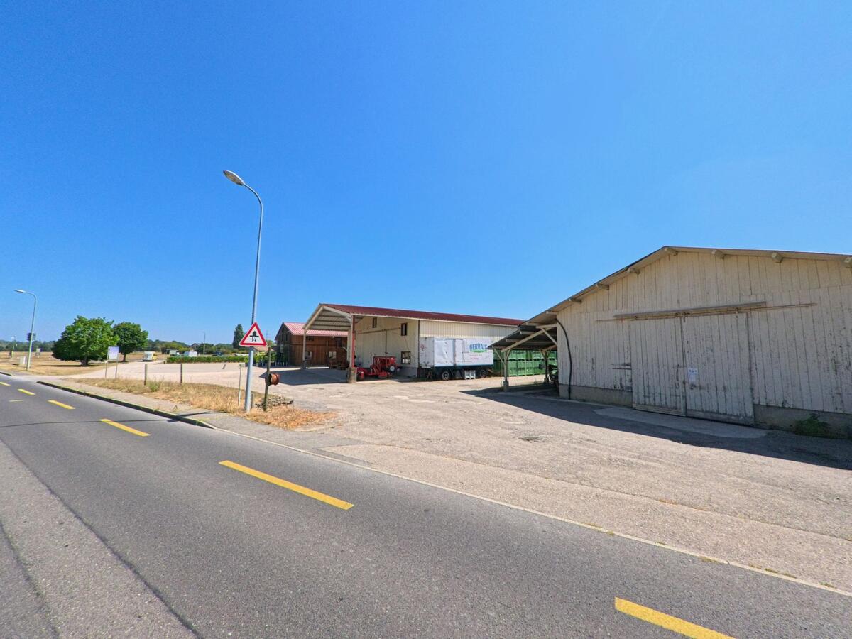 An einer Landstrasse in Russin (1281, Kanton Genf) stehen grosse Wirtschaftsgebäude mit offenen Hallen und landwirtschaftlichen Geräten.