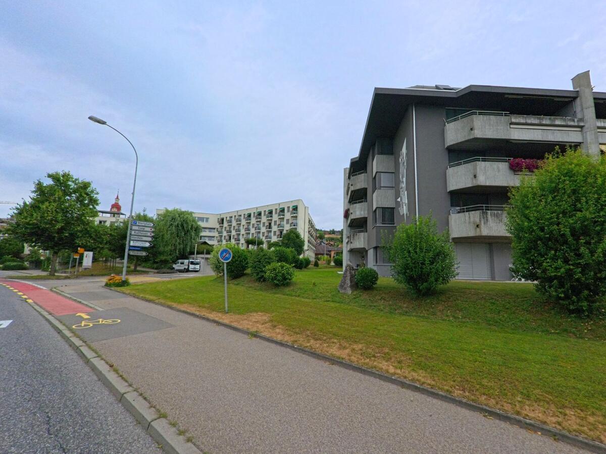In Ruswil (6017, Kanton Luzern) verlaufen ein Fahrradstreifen und ein Gehweg an modernen Wohnbauten vorbei, im Hintergrund ist der Kirchturm des Dorfes zu erkennen.