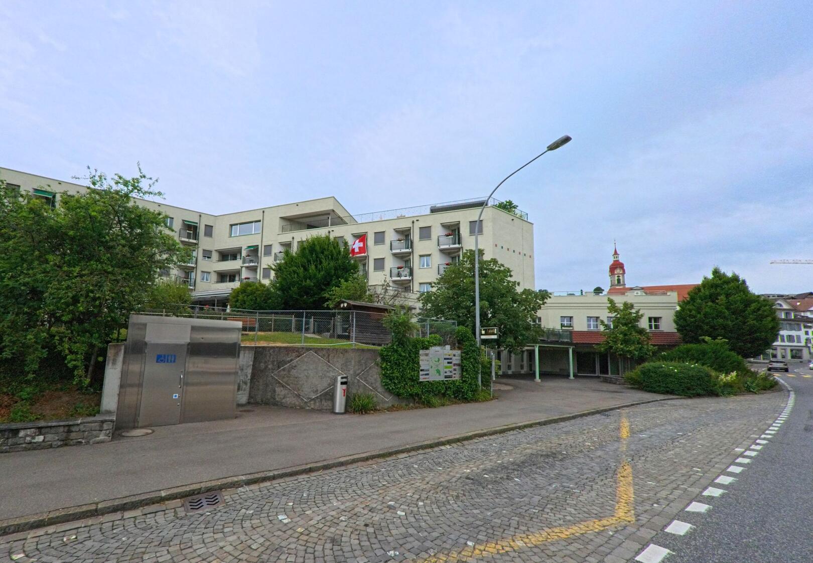 In Ruswil (6017, Kanton Luzern) stehen moderne Wohnbauten mit Balkonen an einer leicht abfallenden Strasse, während im Hintergrund der markante Kirchturm das Dorfbild prägt.