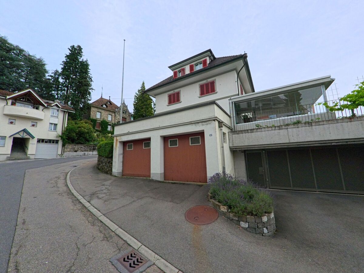 In Ruswil (6017, Kanton Luzern) führt eine kurvige Strasse an Garagen und mehrstöckigen Wohnhäusern vorbei, im Hintergrund ragt ein historisches Gebäude über Bäume hinweg.