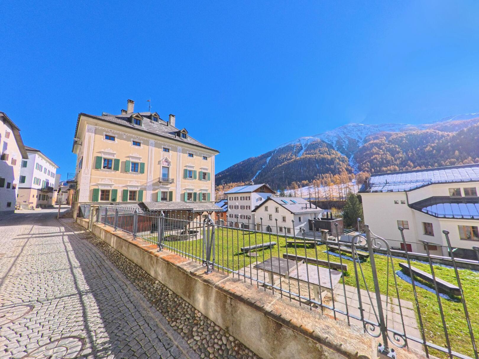 Entlang einer gepflasterten Gasse stehen historische Gebaeude mit Blick auf Gruenflaechen und die alpine Berglandschaft von S-chanf (7525, Kanton Graubuenden) unter klarem Himmel.