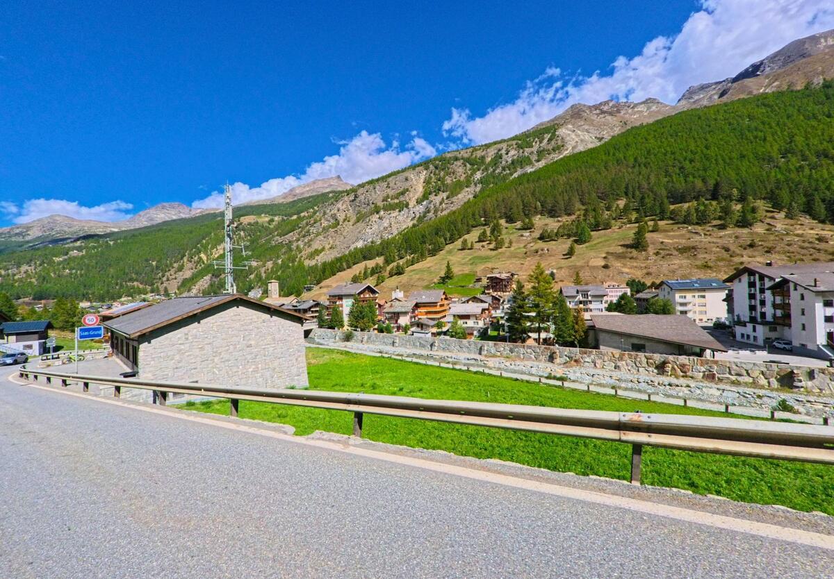 Von der Strasse aus bietet sich ein Blick auf die Siedlung von Saas-Fee (3906, Kanton Wallis) mit Haeusern, gruener Wiese und bewaldeten Berghaengen.