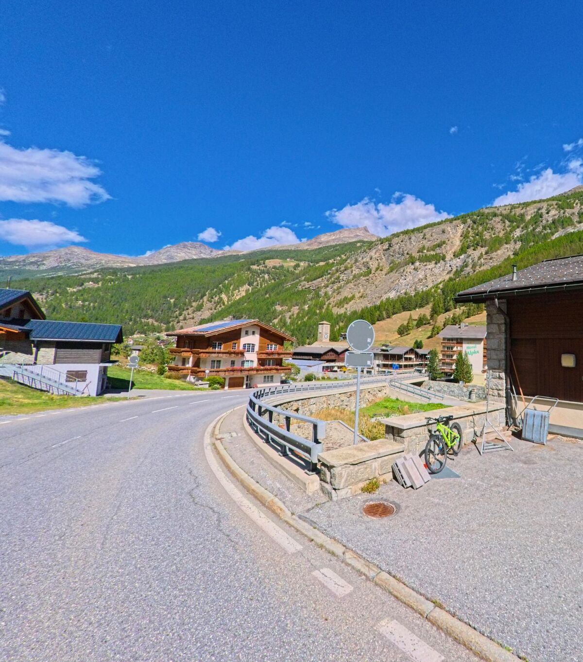 Eine kurvige Strasse fuehrt durch Saas-Fee (3906, Kanton Wallis) vorbei an Chalets und bietet einen Blick auf die umliegenden Berghaenge.