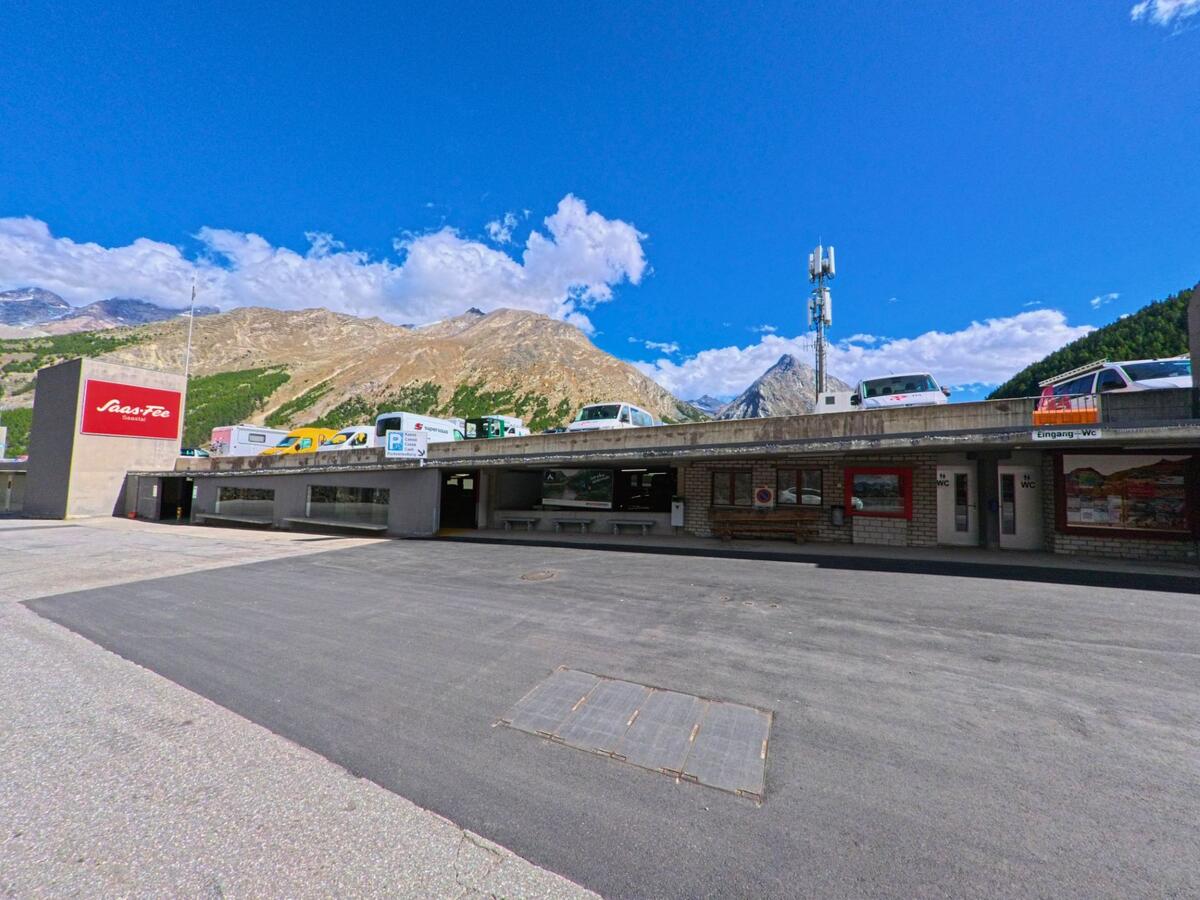 Der Parkplatz mit Beschilderung und Gebaeuden in Saas-Fee (3906, Kanton Wallis) liegt vor einer markanten Bergkulisse unter blauem Himmel.