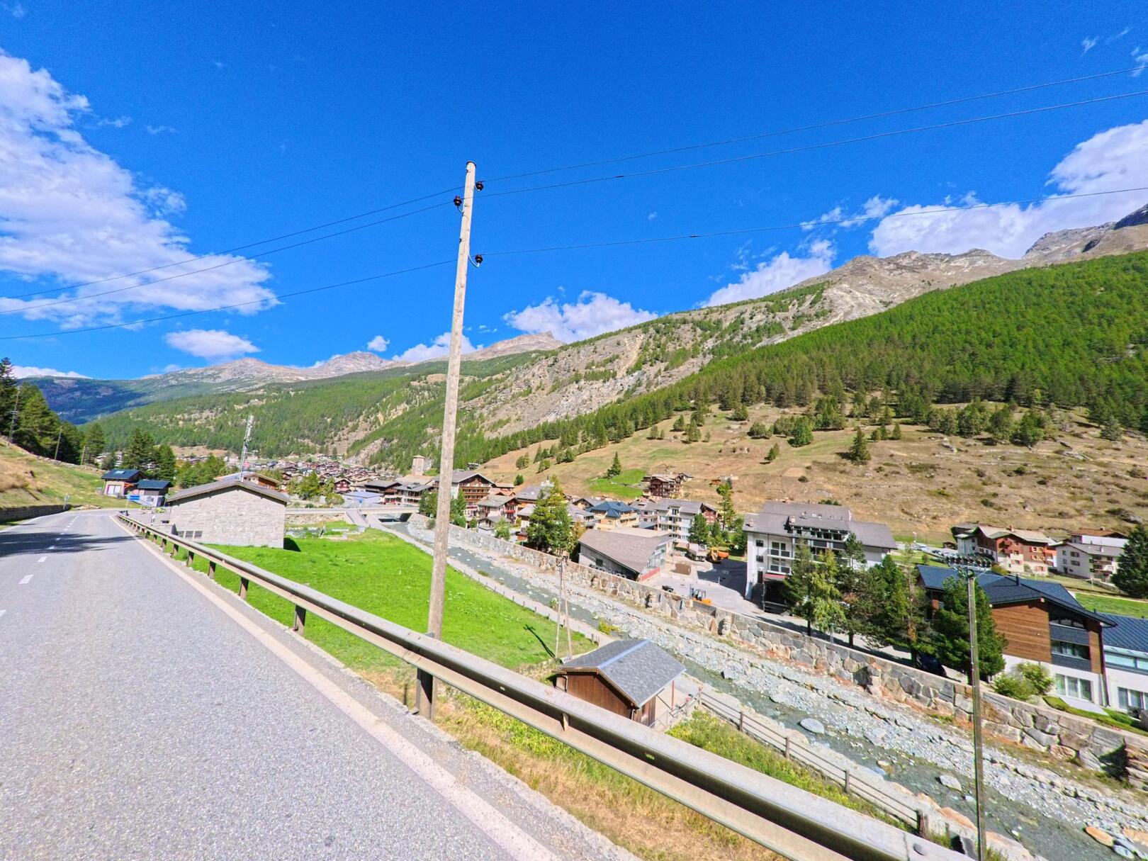 Die Einfahrt nach Saas-Fee (3906, Kanton Wallis) zeigt eine Strasse mit Geländern, flankiert von Wohnhaeusern und Bergen im Hintergrund.