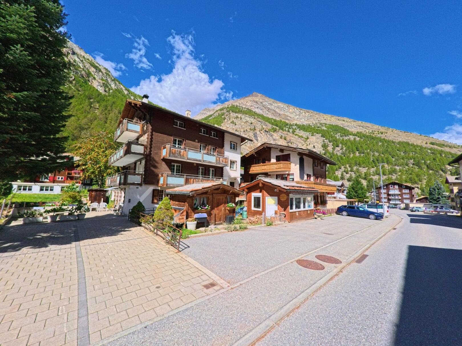 Mehrere traditionelle Holzhäuser in Saas-Almagell (3905, Kanton Wallis) stehen vor einem bewaldeten Berghang unter blauem Himmel.