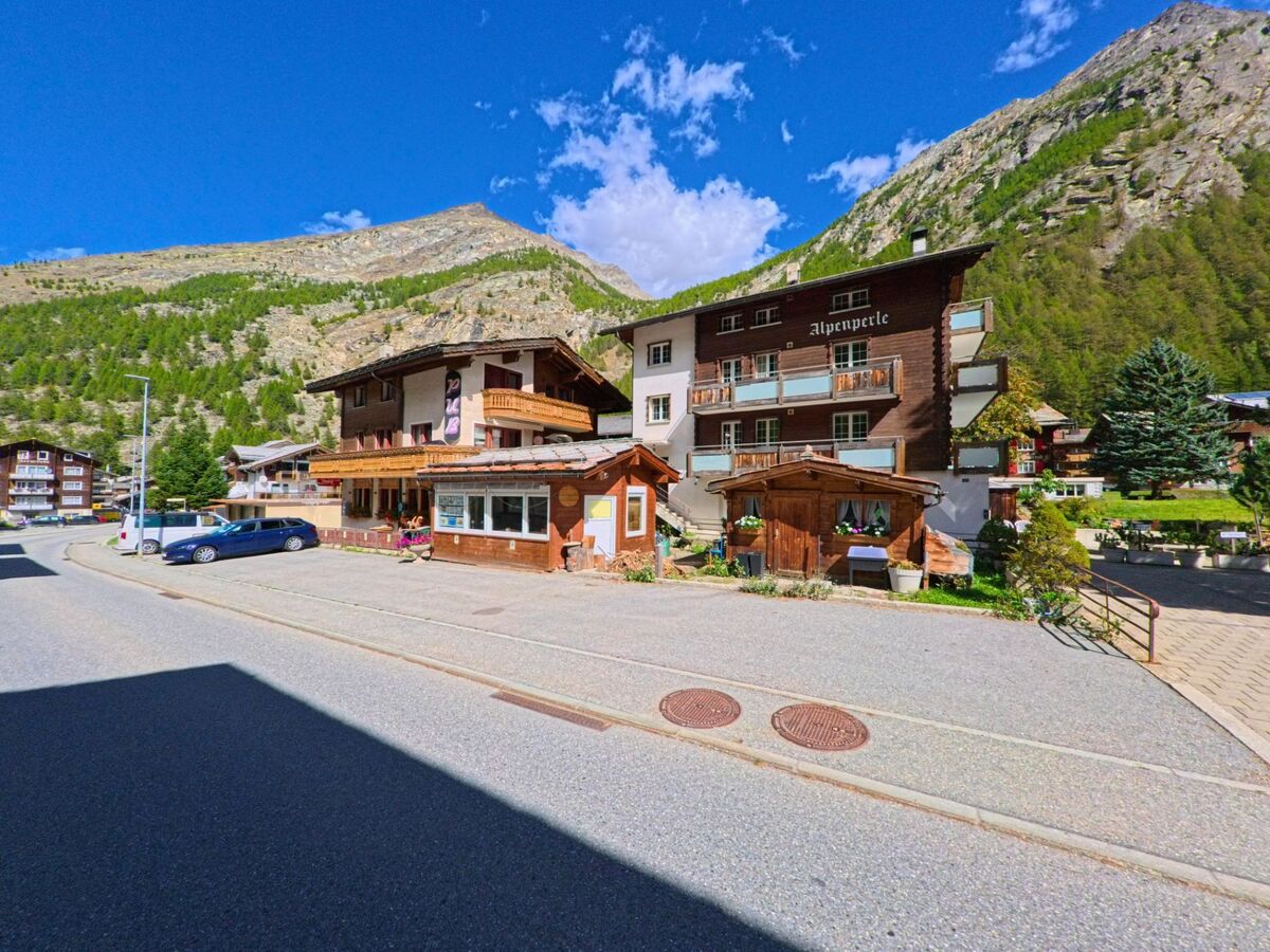 Vor der Bergkulisse in Saas-Almagell (3905, Kanton Wallis) stehen mehrere Gasthaeuser und Hotels mit Holzbalkonen an einer ruhigen Strasse.