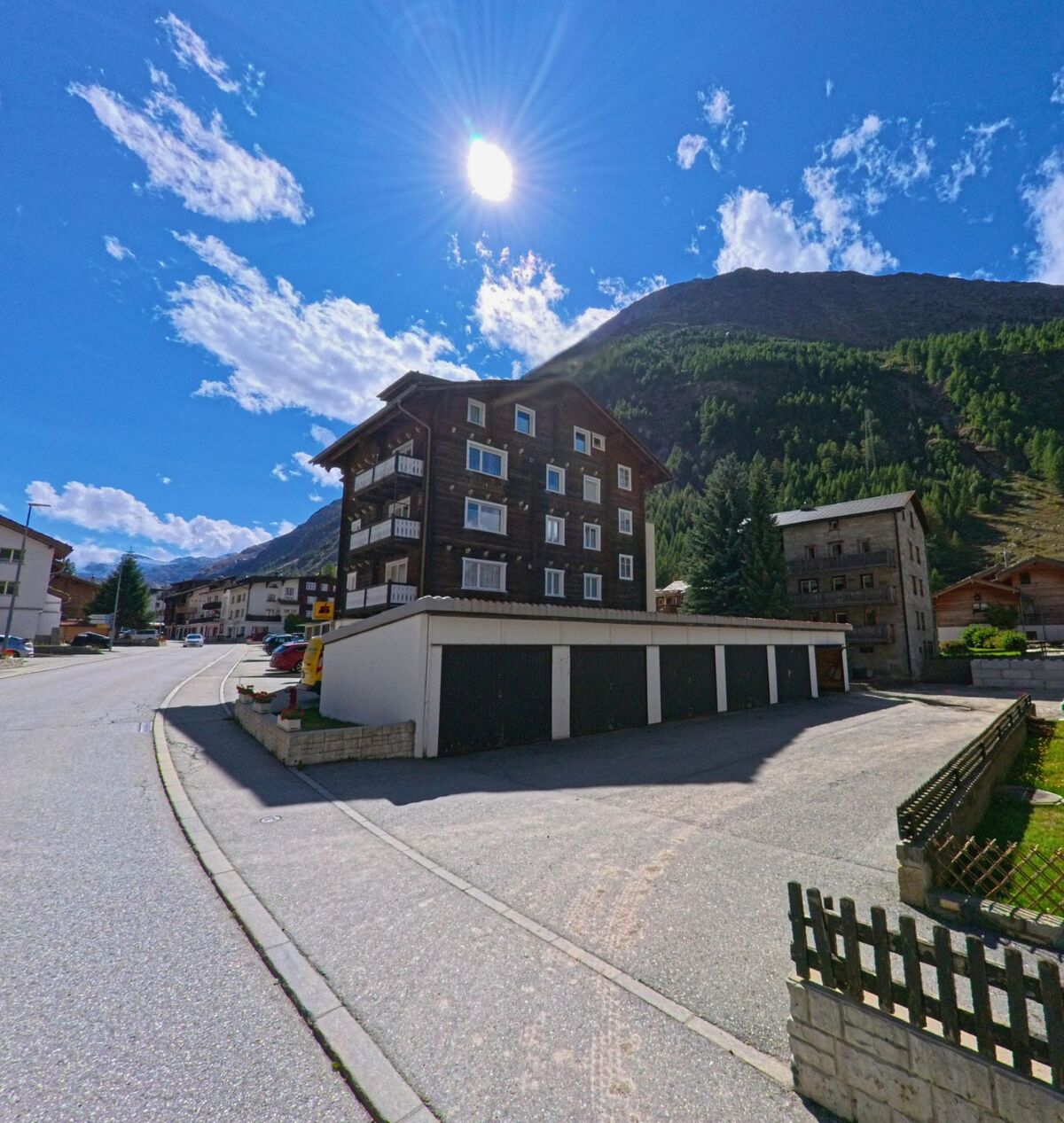 Ein mehrgeschossiges Holzhaus in Saas-Almagell (3905, Kanton Wallis) steht an einer Strasse, waehrend die Sonne ueber den Bergen scheint.