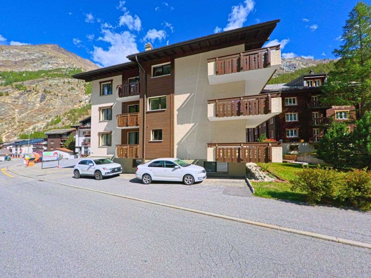 Ein modernes Wohnhaus mit Balkonen und abgestelltem Auto steht in Saas-Almagell (3905, Kanton Wallis) vor einer Kulisse aus Bergen und Waldflaechen.