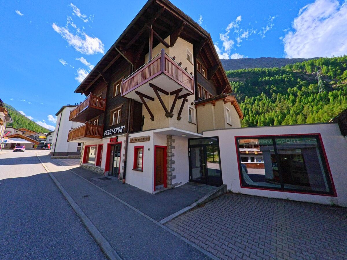 Ein Sportgeschaeft mit Holzbalkonen und angeschlossenem Wohnhaus befindet sich in Saas-Almagell (3905, Kanton Wallis) an einer Strasse mit Blick auf bewaldete Haenge.