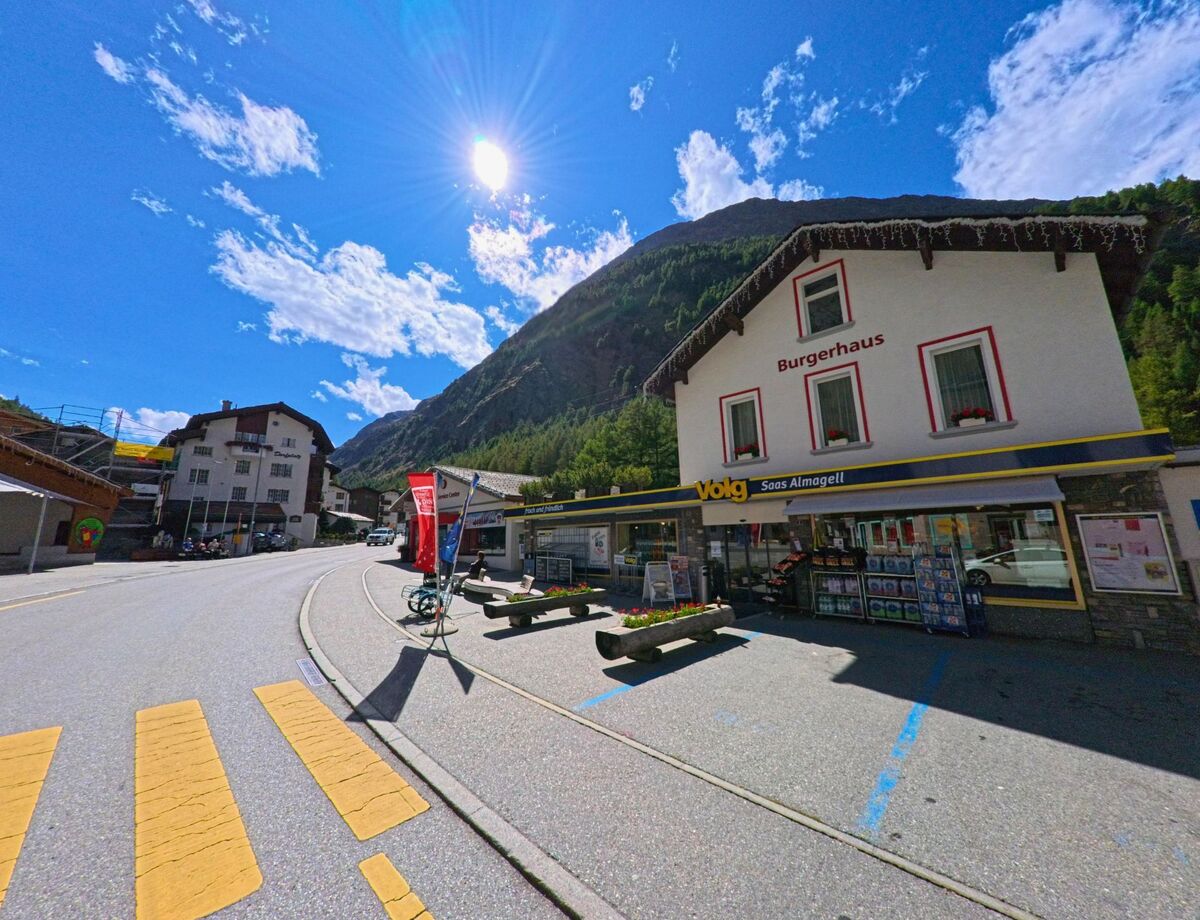 Ein Volg-Laden im Gebaeude Burgerhaus in Saas-Almagell (3905, Kanton Wallis) liegt an der Hauptstrasse mit gelbem Fussgaengerstreifen im Vordergrund.