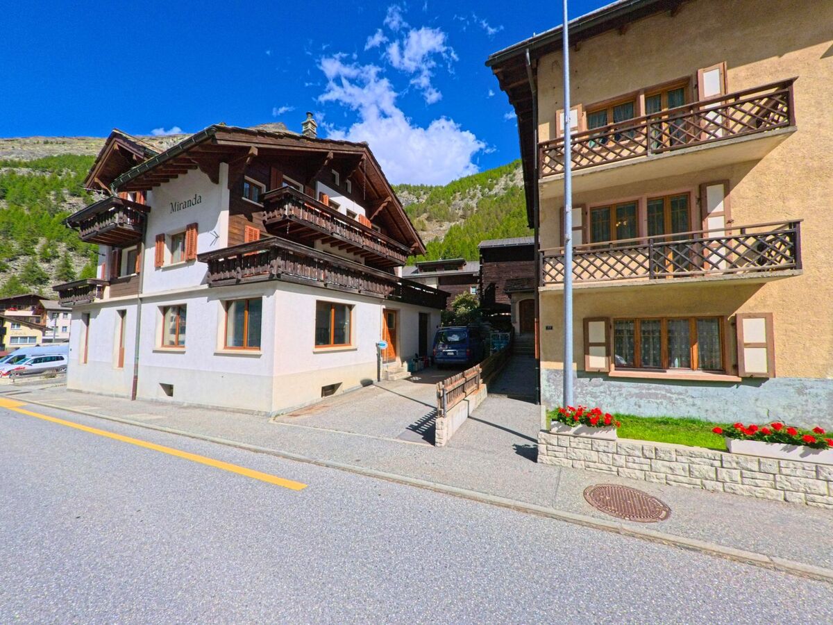 Zwei nebeneinanderliegende Wohnhaeuser mit Holzbalkonen und Blumenschmuck stehen in Saas-Almagell (3905, Kanton Wallis) an einer sonnigen Strasse.