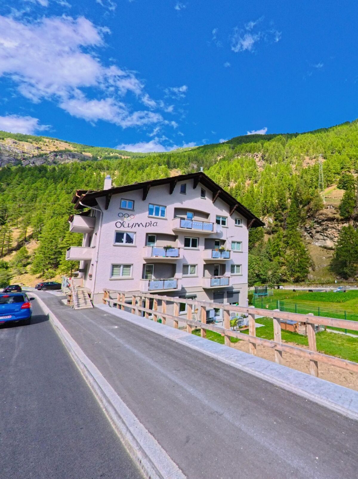Ein mehrstöckiges Wohnhaus mit der Aufschrift Olympia steht in Saas-Balen (3908, Kanton Wallis) am Strassenrand vor einem bewaldeten Hang.