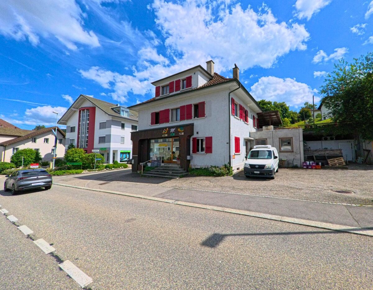 An einer Strassenecke in Safenwil (5745, Kanton Aargau) befindet sich ein kleines Geschäft im Erdgeschoss eines Wohnhauses mit roten Fensterläden, umgeben von weiteren Wohnbauten.