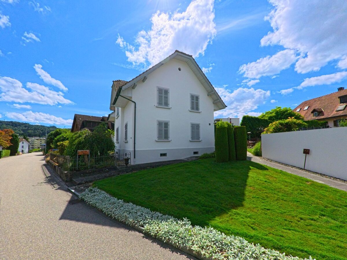 Ein freistehendes Einfamilienhaus mit weissem Anstrich und Fensterläden steht in Safenwil (5745, Kanton Aargau) an einer ruhigen Wohnstrasse mit gepflegtem Vorgarten.