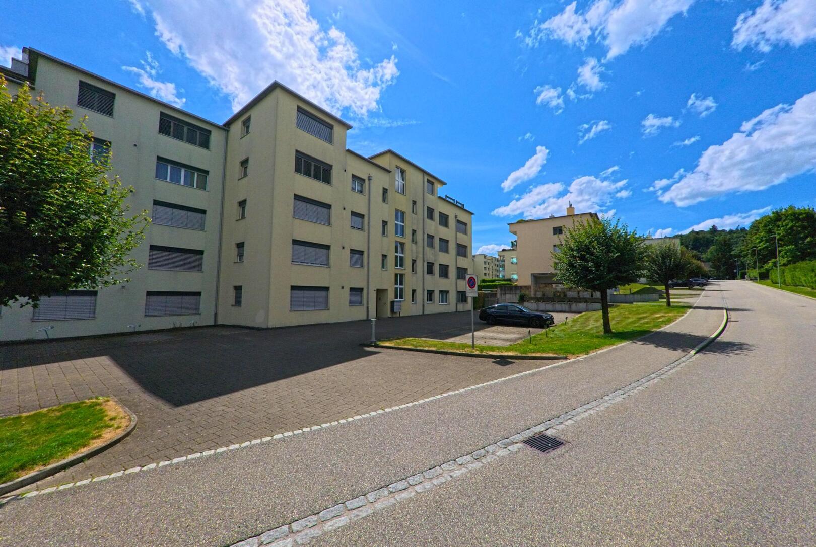 In einem Wohnquartier von Safenwil (5745, Kanton Aargau) reihen sich mehrere mehrstöckige Wohnhäuser entlang einer leicht ansteigenden Strasse mit Baumreihe.